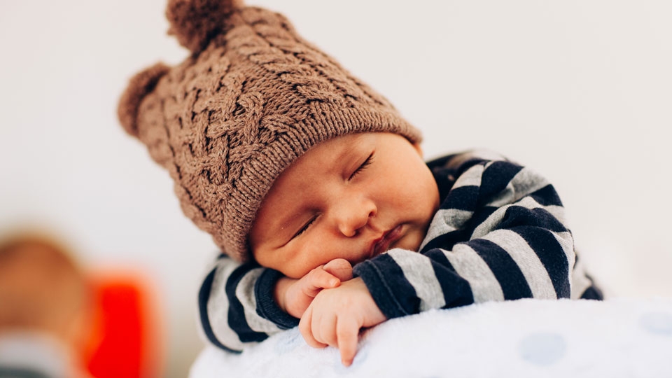 A baby boy sleeping on his arms wearing a warm hat and a striped shirt